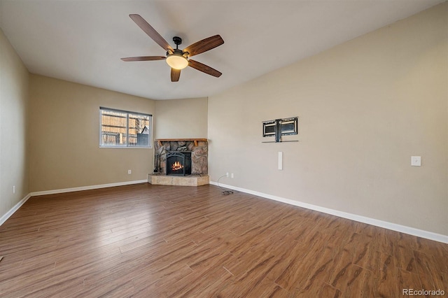 unfurnished living room featuring a stone fireplace, wood finished floors, baseboards, and a ceiling fan