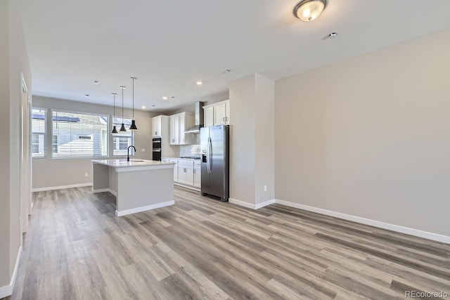 kitchen with decorative light fixtures, sink, stainless steel refrigerator with ice dispenser, a center island with sink, and wall chimney exhaust hood