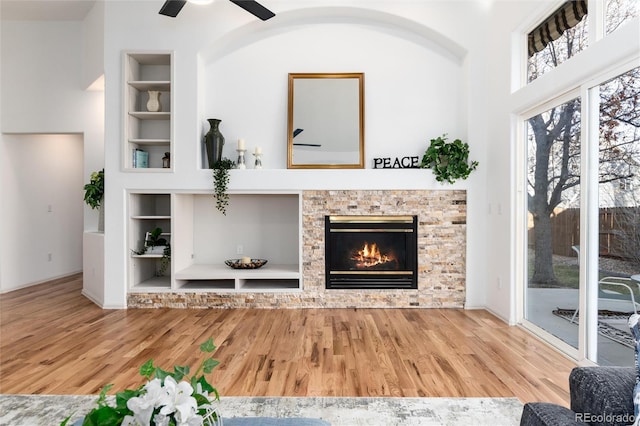 living area with built in shelves, a glass covered fireplace, wood finished floors, baseboards, and ceiling fan