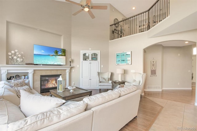 living room featuring a ceiling fan, a high ceiling, a tiled fireplace, visible vents, and arched walkways