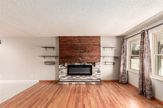 unfurnished living room with a textured ceiling, a stone fireplace, wood finished floors, and baseboards