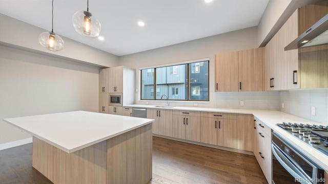 kitchen with appliances with stainless steel finishes, a kitchen island, wall chimney range hood, light brown cabinetry, and hanging light fixtures