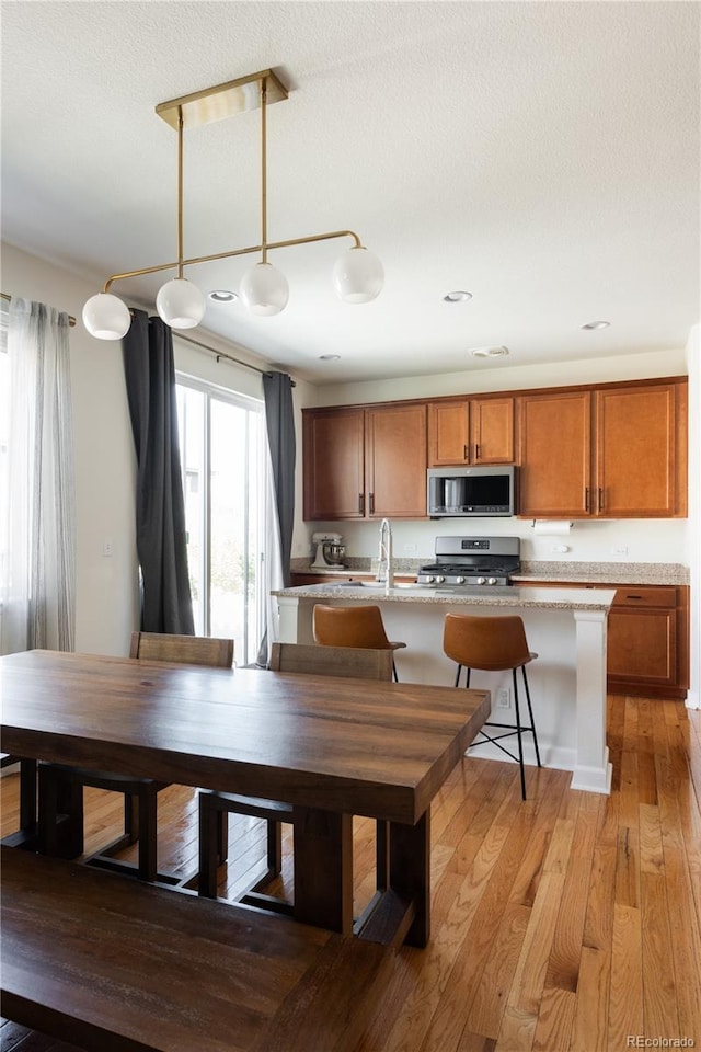 kitchen with brown cabinets, hanging light fixtures, appliances with stainless steel finishes, light wood-style floors, and a kitchen island with sink