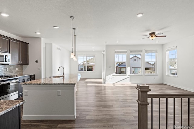 kitchen with sink, a kitchen island with sink, stainless steel appliances, dark brown cabinetry, and light stone countertops