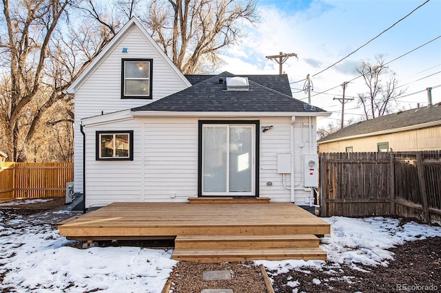 snow covered house featuring a shingled roof, fence, and a deck