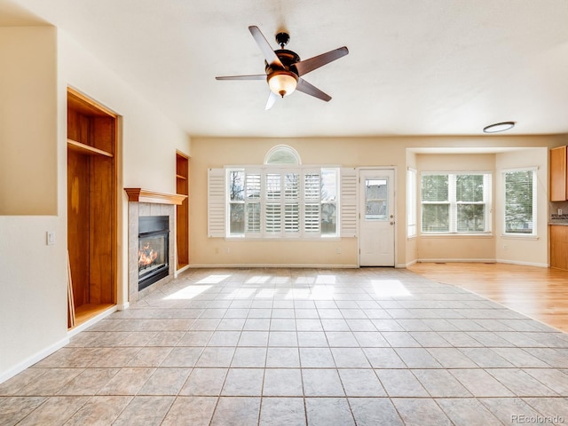 unfurnished living room featuring built in shelves, a tile fireplace, ceiling fan, and baseboards