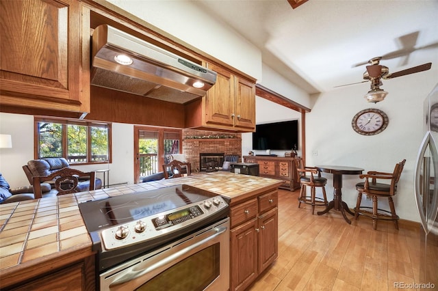 kitchen featuring ceiling fan, appliances with stainless steel finishes, a brick fireplace, brown cabinets, and tile counters
