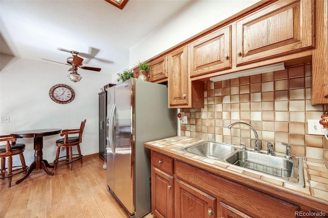 kitchen with brown cabinetry, decorative backsplash, a ceiling fan, stainless steel refrigerator with ice dispenser, and a sink