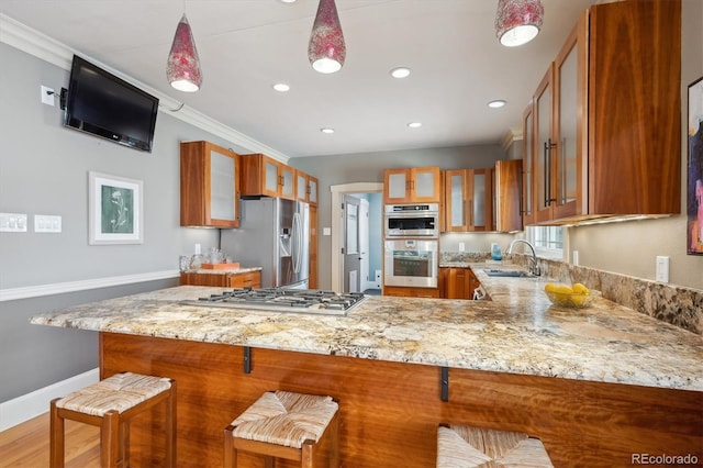 kitchen featuring light stone counters, a sink, appliances with stainless steel finishes, brown cabinetry, and glass insert cabinets