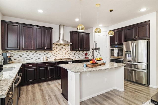 kitchen with a kitchen island, appliances with stainless steel finishes, wall chimney exhaust hood, light stone countertops, and light wood-style flooring