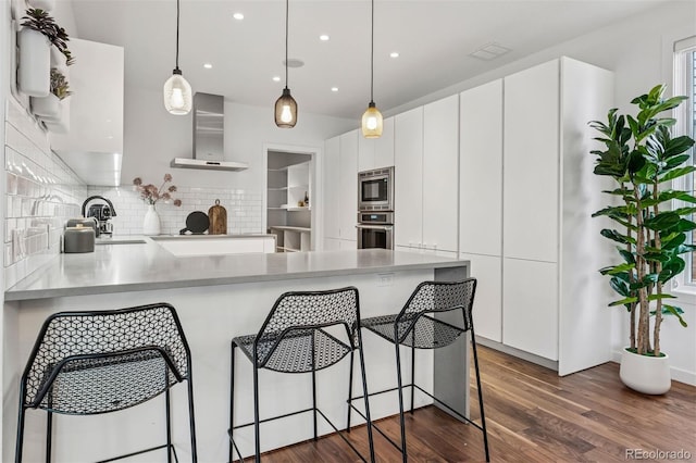 kitchen featuring dark wood-style flooring, a peninsula, wall chimney exhaust hood, backsplash, and stainless steel appliances