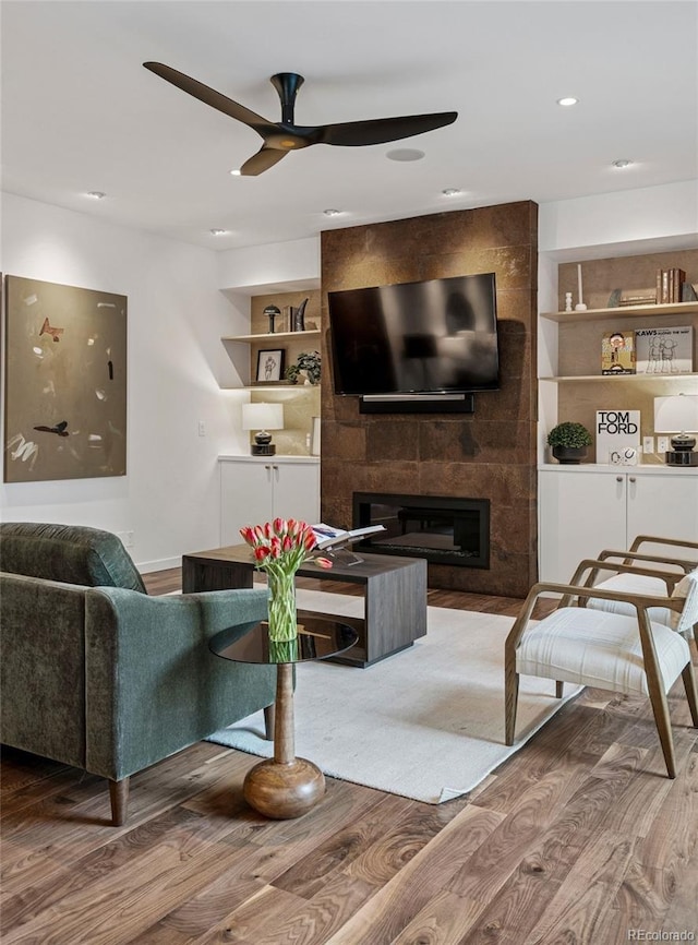 living area with ceiling fan, a large fireplace, built in shelves, wood finished floors, and recessed lighting