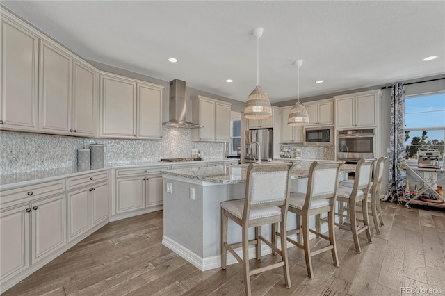 kitchen with appliances with stainless steel finishes, wall chimney exhaust hood, decorative light fixtures, light wood-type flooring, and a kitchen island with sink