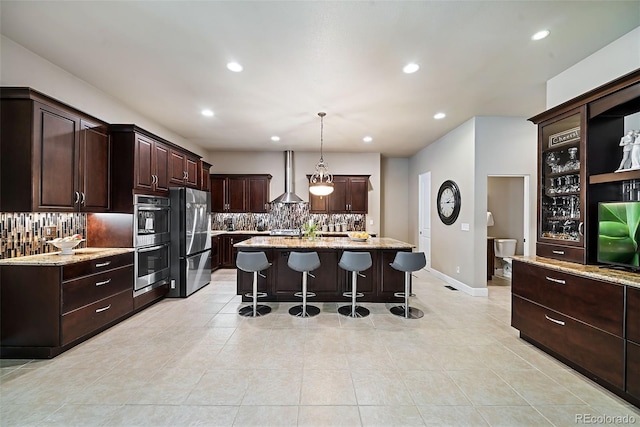 kitchen featuring wall chimney range hood, a breakfast bar area, backsplash, stainless steel appliances, and a center island with sink