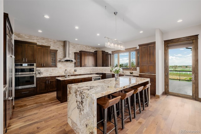 kitchen featuring dark brown cabinetry, double oven, a center island with sink, wall chimney range hood, and backsplash