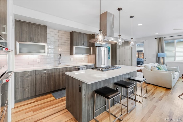 kitchen with appliances with stainless steel finishes, a kitchen breakfast bar, island exhaust hood, light wood-type flooring, and a sink