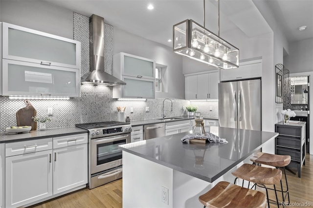 kitchen with dark countertops, wall chimney exhaust hood, appliances with stainless steel finishes, a kitchen breakfast bar, and a sink