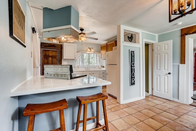 kitchen with a kitchen breakfast bar, ceiling fan, tile counters, white fridge, and white cabinetry