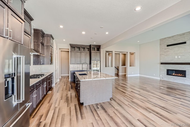 kitchen featuring light hardwood / wood-style floors, a center island with sink, appliances with stainless steel finishes, a fireplace, and dark brown cabinets