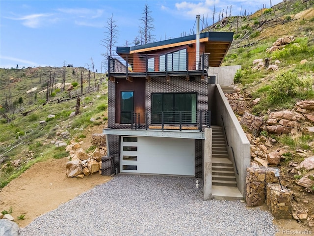 view of front of home featuring a garage, a balcony, stairway, gravel driveway, and brick siding