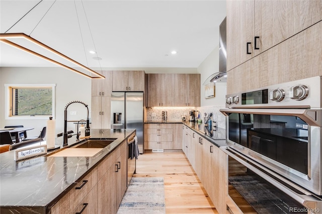 kitchen with a sink, appliances with stainless steel finishes, wall chimney range hood, decorative backsplash, and light brown cabinetry