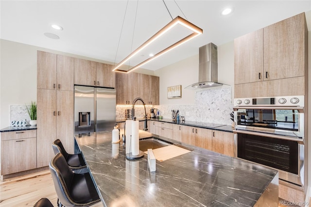 kitchen featuring stainless steel appliances, a sink, wall chimney exhaust hood, and light brown cabinetry