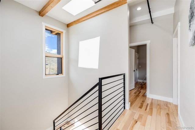 corridor with a skylight, light wood finished floors, baseboards, beamed ceiling, and an upstairs landing