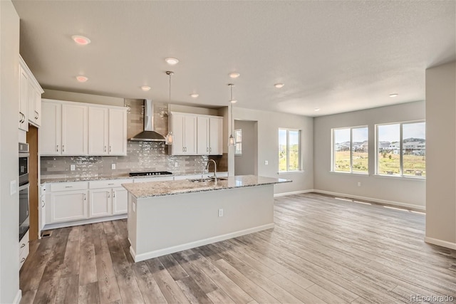 kitchen featuring sink, wall chimney range hood, light hardwood / wood-style flooring, a kitchen island with sink, and white cabinets