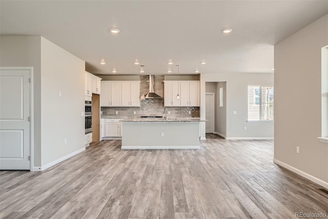 kitchen with a center island with sink, white cabinets, wall chimney range hood, and light stone counters