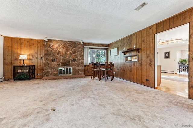 carpeted living area with visible vents, a stone fireplace, wooden walls, a textured ceiling, and a baseboard heating unit
