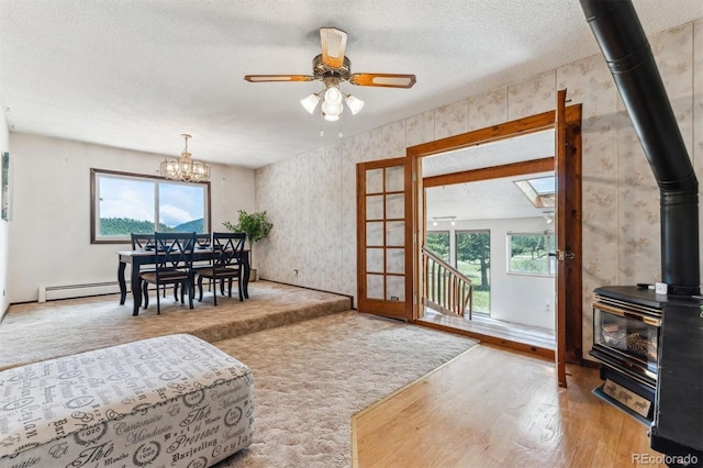 living room with a wood stove, a textured ceiling, ceiling fan with notable chandelier, wood finished floors, and a baseboard heating unit
