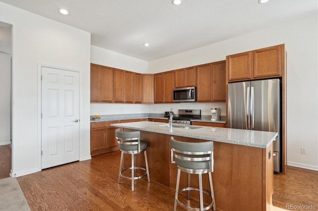 kitchen featuring hardwood / wood-style flooring, appliances with stainless steel finishes, a kitchen island with sink, light stone countertops, and a kitchen bar