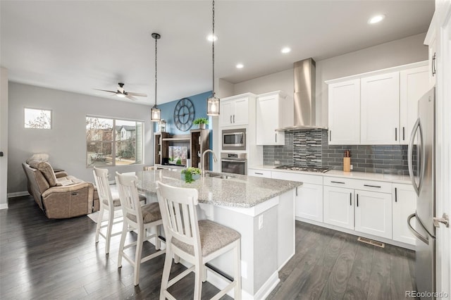 kitchen featuring open floor plan, wall chimney range hood, appliances with stainless steel finishes, dark wood-style floors, and tasteful backsplash