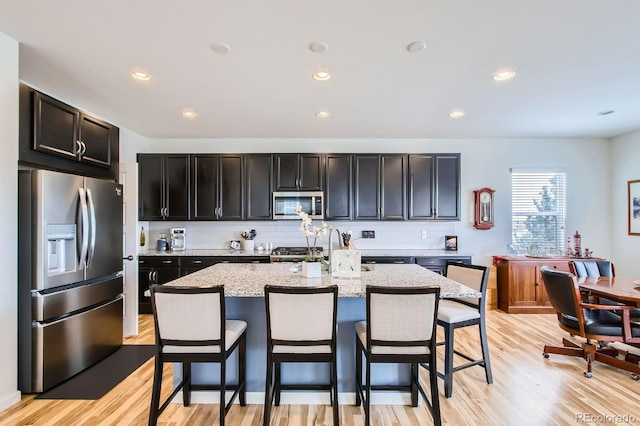 kitchen with light wood finished floors, an island with sink, stainless steel appliances, a kitchen breakfast bar, and decorative backsplash