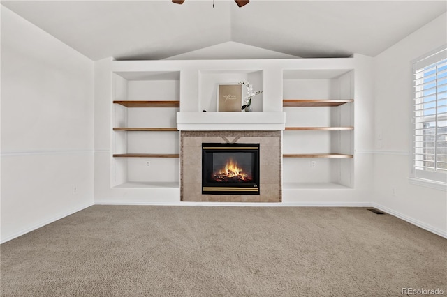 unfurnished living room featuring carpet flooring, built in shelves, vaulted ceiling, ceiling fan, and a tiled fireplace