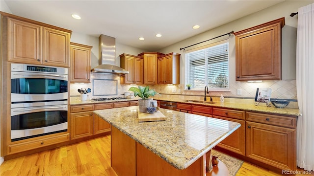 kitchen with wall chimney range hood, sink, stainless steel appliances, a center island, and light stone counters