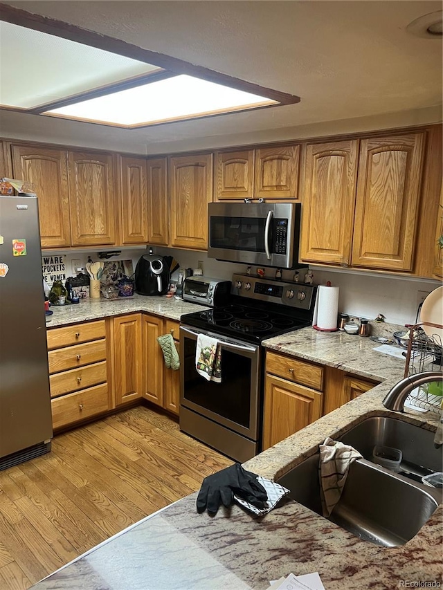kitchen featuring appliances with stainless steel finishes, light wood-type flooring, a sink, light stone counters, and brown cabinets