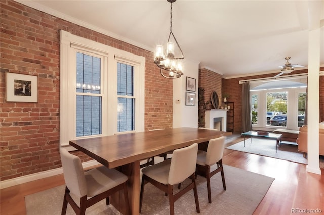 dining area featuring ornamental molding, wood finished floors, brick wall, a large fireplace, and ceiling fan with notable chandelier