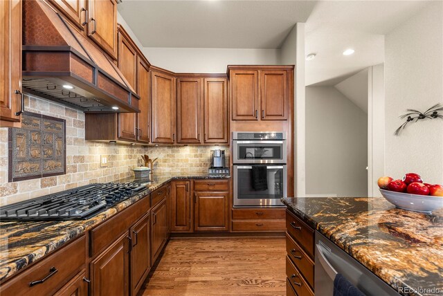 kitchen with wood finished floors, stainless steel appliances, premium range hood, tasteful backsplash, and dark stone countertops