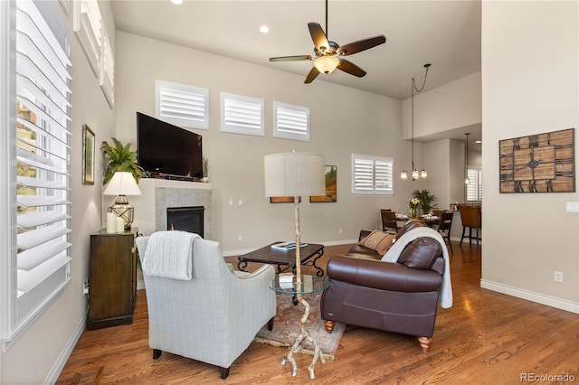 living area with ceiling fan with notable chandelier, baseboards, a towering ceiling, a tile fireplace, and wood finished floors