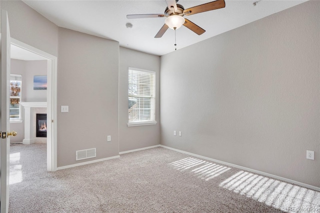 empty room with carpet flooring, visible vents, baseboards, a ceiling fan, and a glass covered fireplace