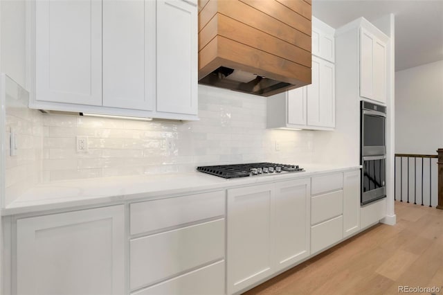 kitchen featuring light stone counters, custom range hood, stainless steel appliances, light hardwood / wood-style floors, and white cabinets