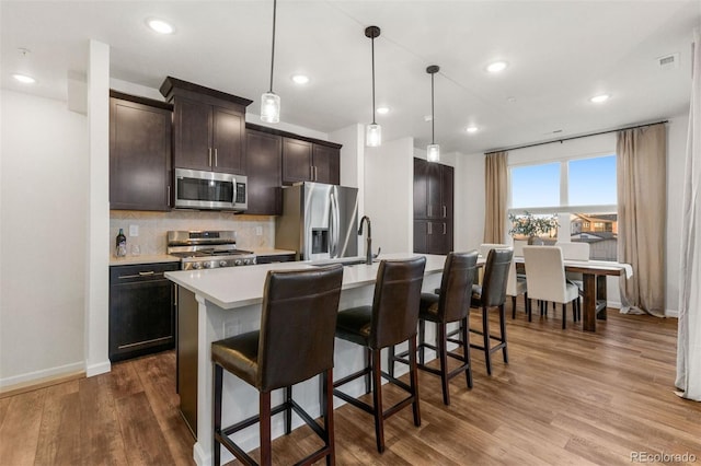 kitchen with appliances with stainless steel finishes, decorative light fixtures, backsplash, hardwood / wood-style flooring, and a center island with sink