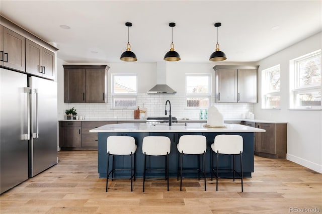 kitchen with pendant lighting, built in refrigerator, wall chimney exhaust hood, an island with sink, and dark brown cabinets