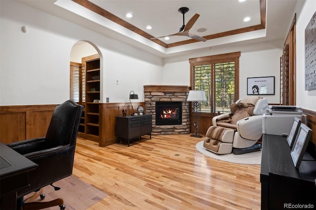 home office with light wood finished floors, ceiling fan, a tray ceiling, and a stone fireplace