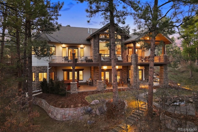 back of house at dusk with stone siding, stucco siding, french doors, and a balcony