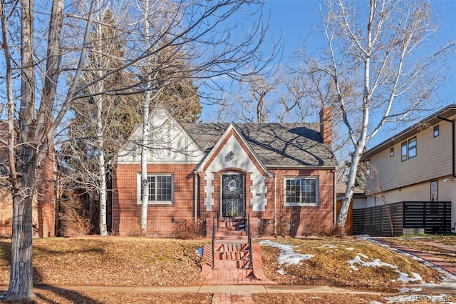 tudor home with a shingled roof, brick siding, fence, and a chimney