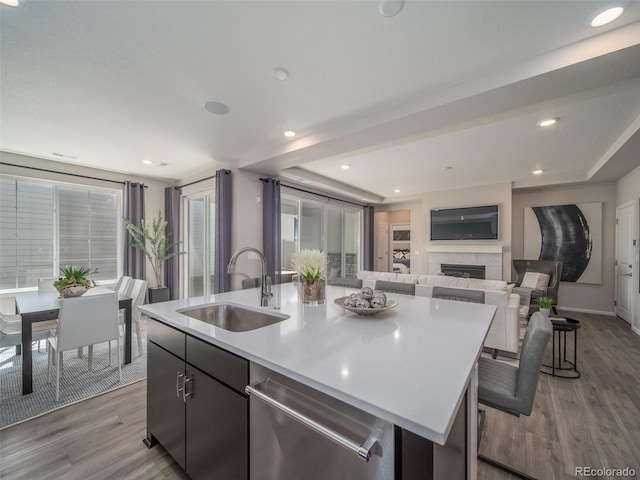 kitchen featuring a center island with sink, sink, stainless steel dishwasher, light wood-type flooring, and a tiled fireplace