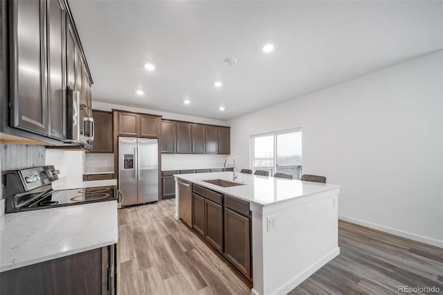 kitchen with a kitchen island with sink, light wood-style flooring, dark brown cabinetry, stainless steel appliances, and a sink