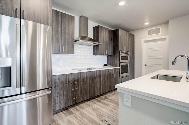 kitchen with stainless steel appliances, wall chimney range hood, decorative backsplash, sink, and light hardwood / wood-style floors